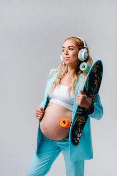 A pregnant girl in a turquoise suit with a skateboard in her hands and headphones stands on a gray background.