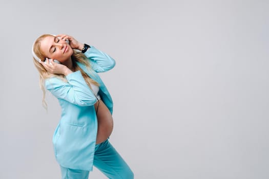 a pregnant woman in a turquoise suit with headphones stands and listens to music on a gray background.