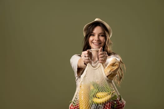 Brunette woman in beige t-shirt hat holding cotton reusable mesh shopping bags with vegetables, groceries and looks in a camera on pastel green background. Zero waste, plastic free concept