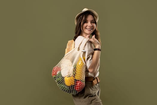 Smiling young woman in light summer clothes with a mesh eco bag full of vegetables, greens on a green studio background. Sustainable lifestyle. Eco friendly concept. Zero waste