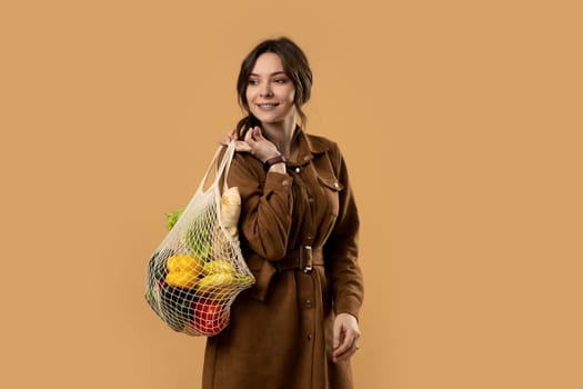 Portrait of happy smiling young woman in brown dress holding reusable string bag with groceries over orange background. Sustainability, eco living and people concept