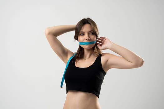Woman in a black top with a blue measuring tape over a mouth, white background. Healthy lifestyle concept. Diet, weight loss