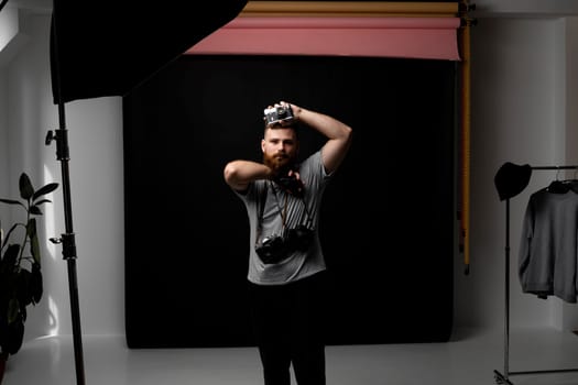 Bearded professional photographer in a grey t-shirt with a bunch of different cameras in a hands and on a shoulder