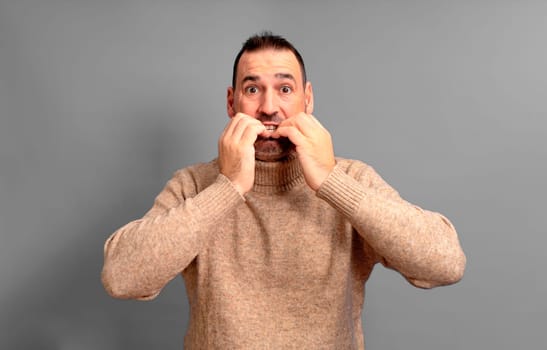 Hispanic man in his 40s scared and shocked biting his nails, staring wide-eyed at something horrible, wearing a turtleneck, standing over gray background, copy space