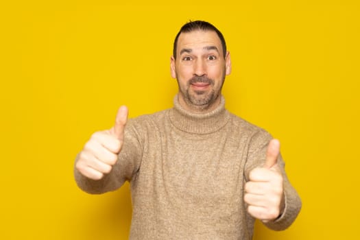 Bearded hispanic man in friendly attitude wearing a beige turtleneck posing funny with thumbs up isolated over yellow background