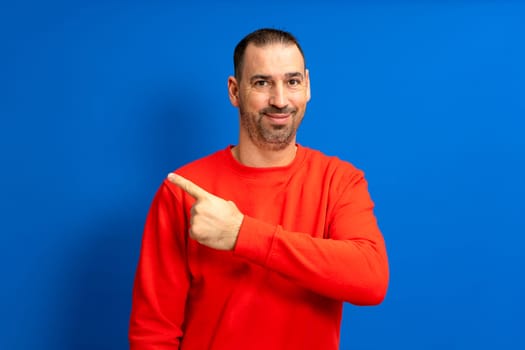 Bearded hispanic man wearing a red sweater smiling at camera while pointing to the side isolated over blue background, indicating a path or direction to follow