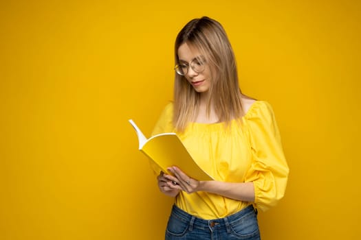 Focused young teenage woman wearing glasses is reading an interesting book isolated over yellow background. Girl reading novel. Studying