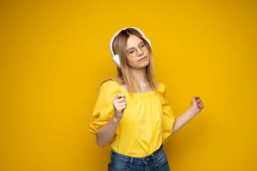Portrait of charming blonde woman in a yellow t-shirt and glasses wearing white wireless headphones and listening to music isolated on yellow background