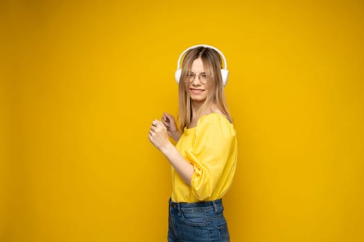 Portrait of happy blonde young woman in a yellow t-shirt and glasses listening a music or podcast with a big white wireless headphones