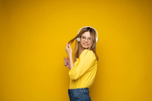 Beautiful attractive young blond woman wearing yellow t-shirt and glasses in white headphones listening music, dancing and laughing on blue background in studio. Relaxing and enjoying. Lifestyle