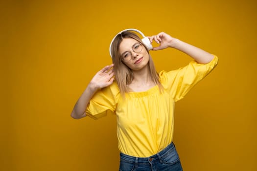 Pretty girl in a yellow t-shirt and glasses listening music with her headphones and dancing on yellow background