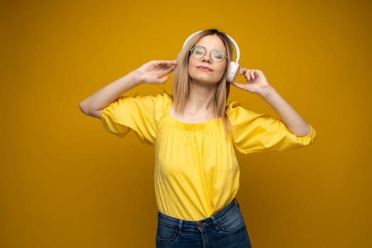 Beautiful attractive young blond woman wearing yellow t-shirt and glasses in white headphones listening music, dancing and laughing on blue background in studio. Relaxing and enjoying. Lifestyle