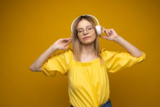 Beautiful attractive young blond woman wearing yellow t-shirt and glasses in white headphones listening music, dancing and laughing on blue background in studio. Relaxing and enjoying. Lifestyle