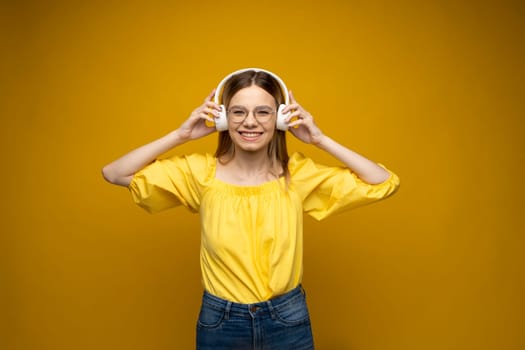 Beautiful attractive young blond woman wearing yellow t-shirt and glasses in white headphones listening music, dancing and laughing on blue background in studio. Relaxing and enjoying. Lifestyle