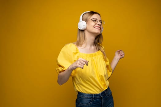 Cheerful young pretty girl smiling while listening music in headphones and dancing on yellow background