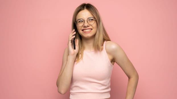 Closeup photo of attractive positive laughing young blonde woman wearing stylish yellow shirt and glasses communicating on mobile phone and smiling isolated over yellow background