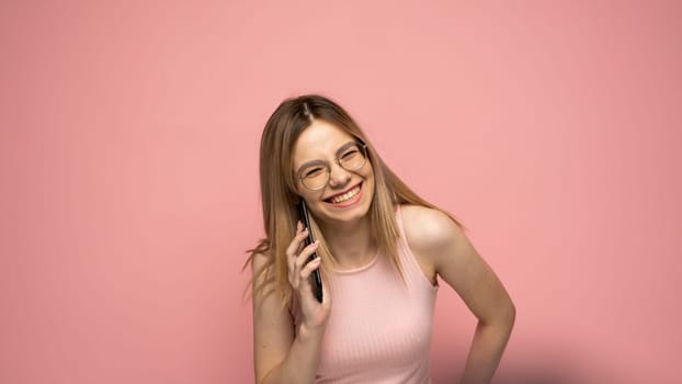 Closeup photo of attractive positive laughing young blonde woman wearing stylish yellow shirt and glasses communicating on mobile phone and smiling isolated over yellow background