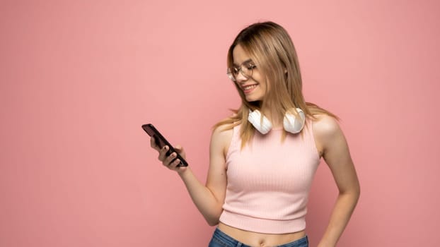 Surprised young happy girl in pink t-shirt using mobile phone, typing sms message isolated on pink background, studio portrait. People lifestyle concept. Mock up copy space