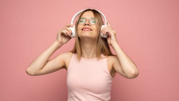 Beautiful attractive young blond woman wearing pink t-shirt and glasses in white headphones listening music and smiling on pink background in studio. Relaxing and enjoying. Lifestyle