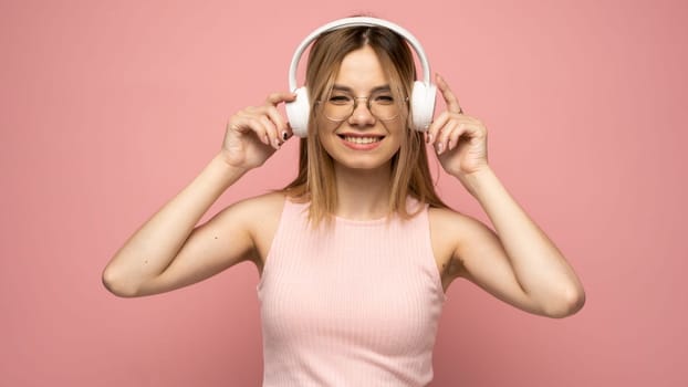 Beautiful attractive young blond woman wearing pink t-shirt and glasses in white headphones listening music and smiling on pink background in studio. Relaxing and enjoying. Lifestyle