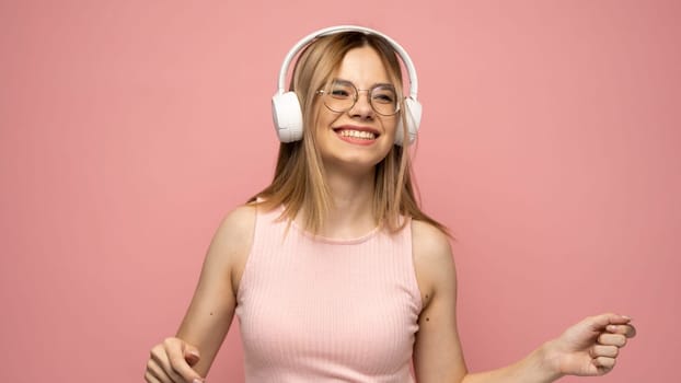 Pretty girl in a pink t-shirt and glasses listening music with her headphones and dancing on pink background