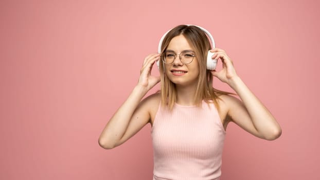 Beautiful young blonde woman with headphones and mobile device listening to music and smiling and dancing, isolated on pink background