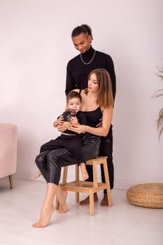 A stylish multi-racial family, with a little boy, in black clothes, sitting in the room. Vertical