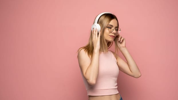 Happy girl in a glasses and pink shirt dancing and listening to the music isolated on a pink background