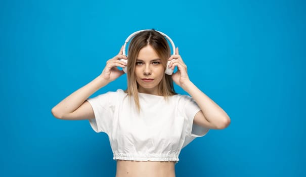 Beautiful attractive young blond woman wearing white t-shirt and glasses in white headphones listening music and smiling on blue background in studio. Relaxing and enjoying. Lifestyle