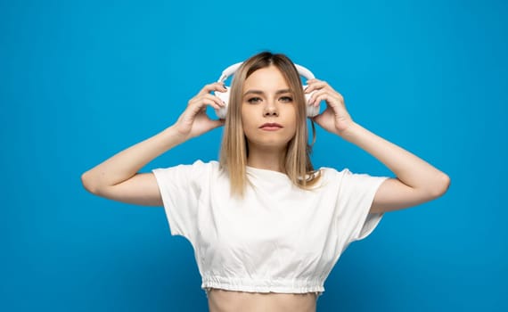 Beautiful attractive young blond woman wearing white t-shirt and glasses in white headphones listening music and smiling on blue background in studio. Relaxing and enjoying. Lifestyle