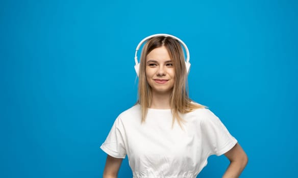 Beautiful attractive young blond woman wearing white t-shirt and glasses in white headphones listening music and smiling on blue background in studio. Relaxing and enjoying. Lifestyle