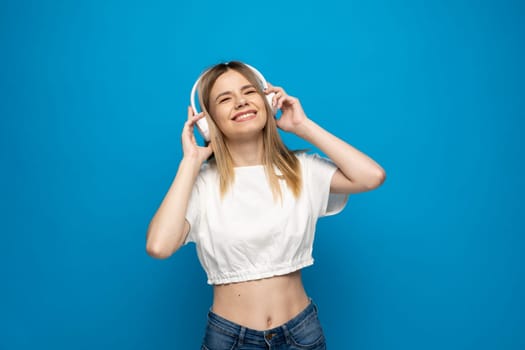 Beautiful attractive young blond woman wearing white t-shirt and glasses in white headphones listening music and dancing on blue background in studio. Relaxing and enjoying. Lifestyle