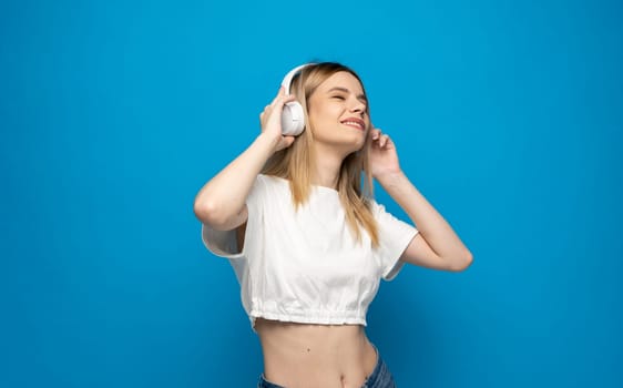 Smiling blonde young woman in a glasses and in headphones listening to music and dancing on blue background