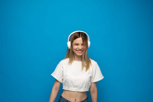 Smiling attractive young blond woman wearing white t-shirt and glasses in white headphones listening music and dancing on blue background in studio. Relaxing and enjoying. Lifestyle