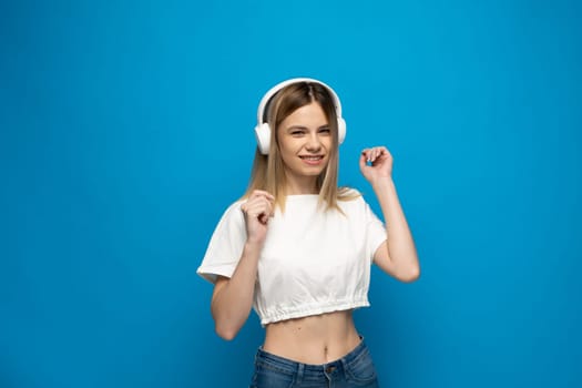 Cheerful young pretty girl smiling while listening music in headphones and dancing on blue background