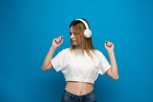 Beautiful young woman in headphones listening to music and dancing on blue background. Young beautiful woman in bright outfit enjoying a music