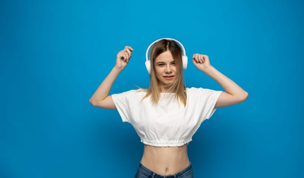 Beautiful attractive young blond woman wearing white t-shirt and glasses in white headphones listening music and dancing on blue background in studio. Relaxing and enjoying. Lifestyle