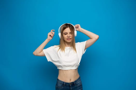 Beautiful attractive young blond woman wearing white t-shirt and glasses in white headphones listening music and dancing on blue background in studio. Relaxing and enjoying. Lifestyle