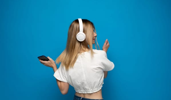 Beautiful young blonde woman with headphones and mobile device listening to music and smiling and dancing, isolated on blue background