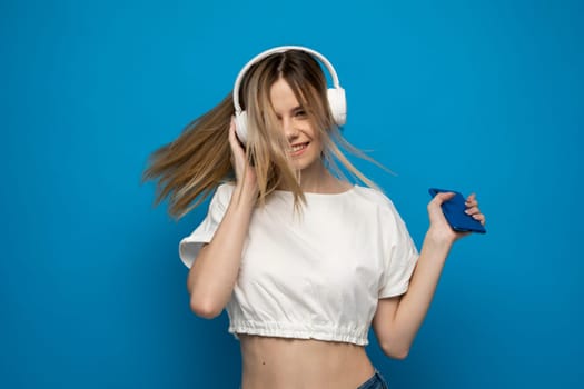 Beautiful attractive young blond woman wearing white t-shirt and glasses in white headphones listening music from smartphone, dancing and laughing on blue background in studio. Lifestyle