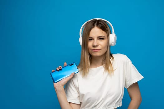 Beautiful young blonde woman with headphones and mobile device listening to music and smiling and dancing, isolated on blue background