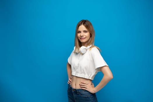 Portraite of young blonde beautiful girl in white outfit with a big headphones on a shoulder on blue background