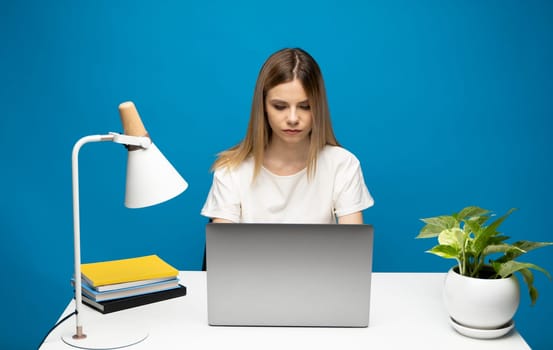 Portrait of a pretty young woman studying while sitting at the table with grey laptop computer, notebook. Smiling business woman working with a laptop isolated on a blue background