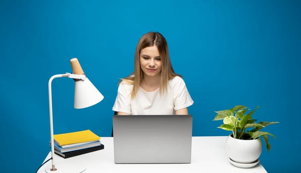 Portrait of a pretty young woman studying while sitting at the table with grey laptop computer, notebook. Smiling business woman working with a laptop isolated on a blue background