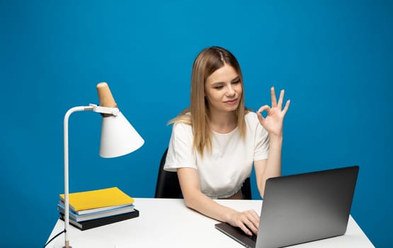 It's okay. Overjoyed woman in a white shirt and glasses showing ok sign, sitting with laptop over blue background
