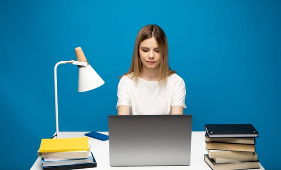 Portrait of a pretty young woman studying while sitting at the table with grey laptop computer, notebook. Smiling business woman working with a laptop isolated on a blue background