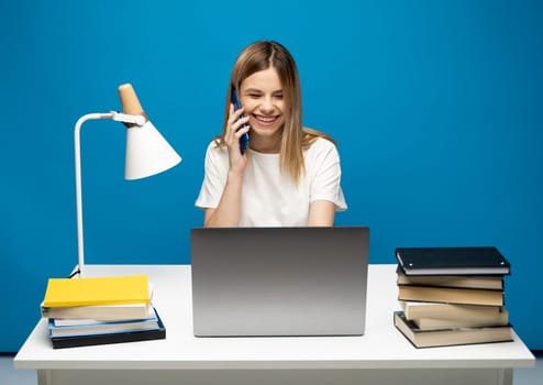 Young attractive business woman with a blond hair sitting at the table and working on a laptop and talking with a client on the phone. Young cheerful student girl talking with a smartphone. Studying