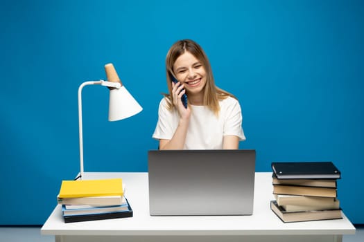 Young attractive business woman with a blond hair sitting at the table and working on a laptop and talking with a client on the phone. Young cheerful student girl talking with a smartphone. Studying