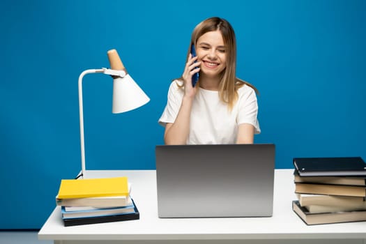 Young attractive business woman with a blond hair sitting at the table and working on a laptop and talking with a client on the phone. Young cheerful student girl talking with a smartphone. Studying