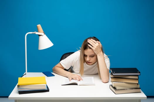 Young student woman in white casual clothes reading the book in library of university or college. Sitting and reading on blue background. Studying